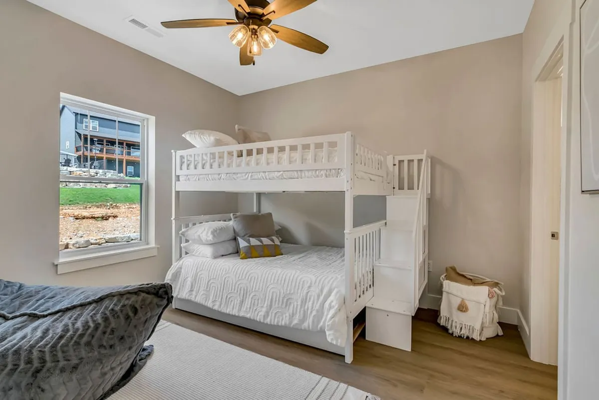 A cozy bunk room featuring a sturdy wooden bunk bed beside a sunlit window, a ceiling fan for comfort, and soft, neutral-colored bedding. The intimate space feels inviting for children or friends to read, play, or rest.