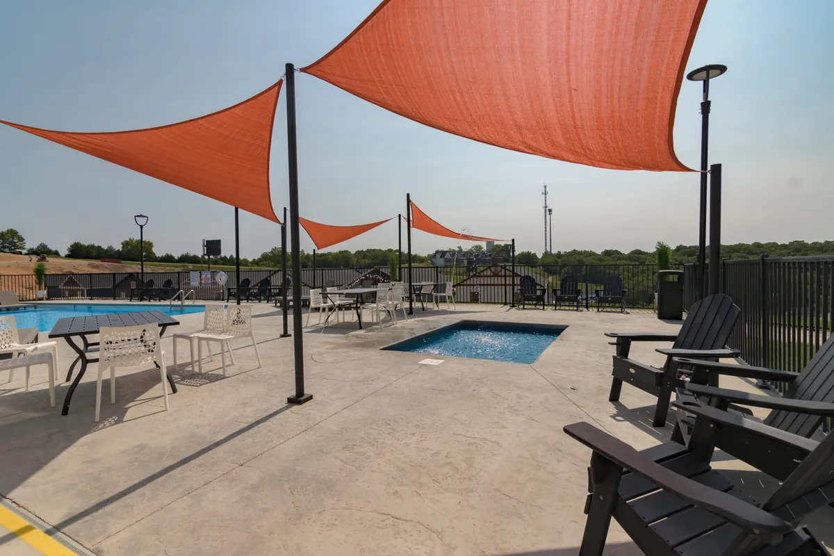 This image features a sparkling outdoor swimming pool surrounded by orange shade sails, lounge chairs, and a seating area with a table. The modern pool area is set against a backdrop of lush greenery, inviting guests to relax, swim, and socialize in the open air.