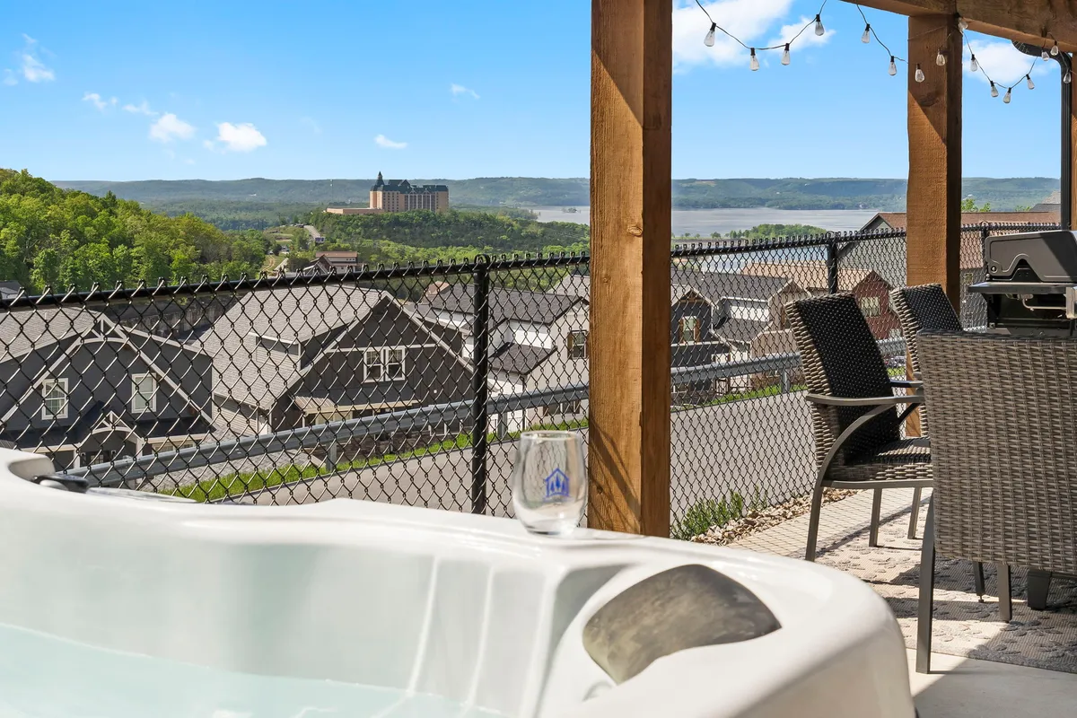 This exterior image captures a private deck featuring a hot tub, an inviting outdoor dining area, string lights overhead, and a stunning lake view in the background. The space is styled for socializing and relaxing, with comfortable seating and an atmosphere perfect for sunset or stargazing.