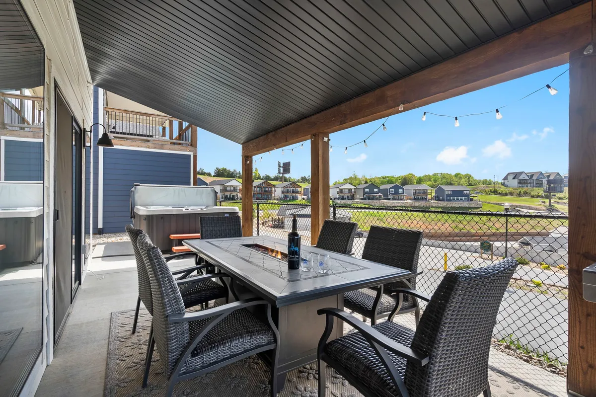 A stylish outdoor living space featuring a dining table, comfortable seating, twinkling string lights, and a private hot tub. The area overlooks neighboring homes and distant views, ideal for gatherings or unwinding after an active day.