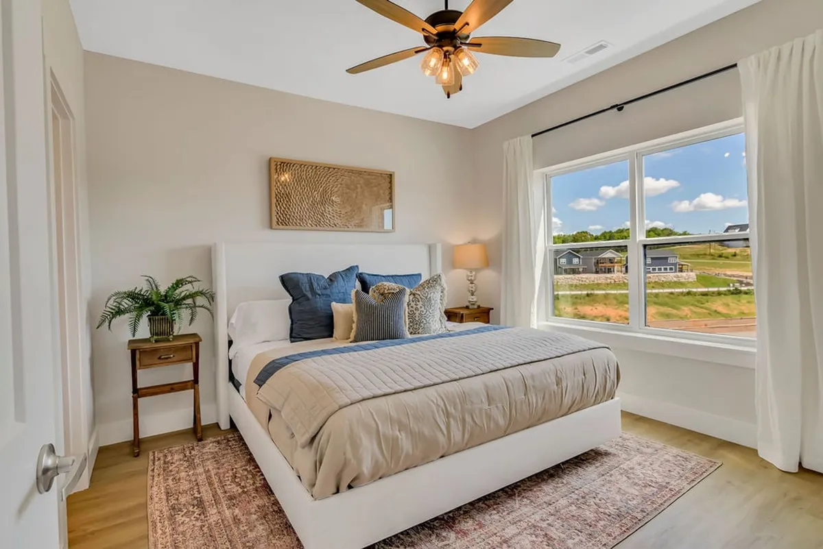 A serene bedroom with a neatly made bed adorned with decorative pillows, a large window that lets in abundant natural light, a nightstand with a lamp, and tasteful wall art. The calm color palette and plush linens create a soothing atmosphere ideal for relaxation.