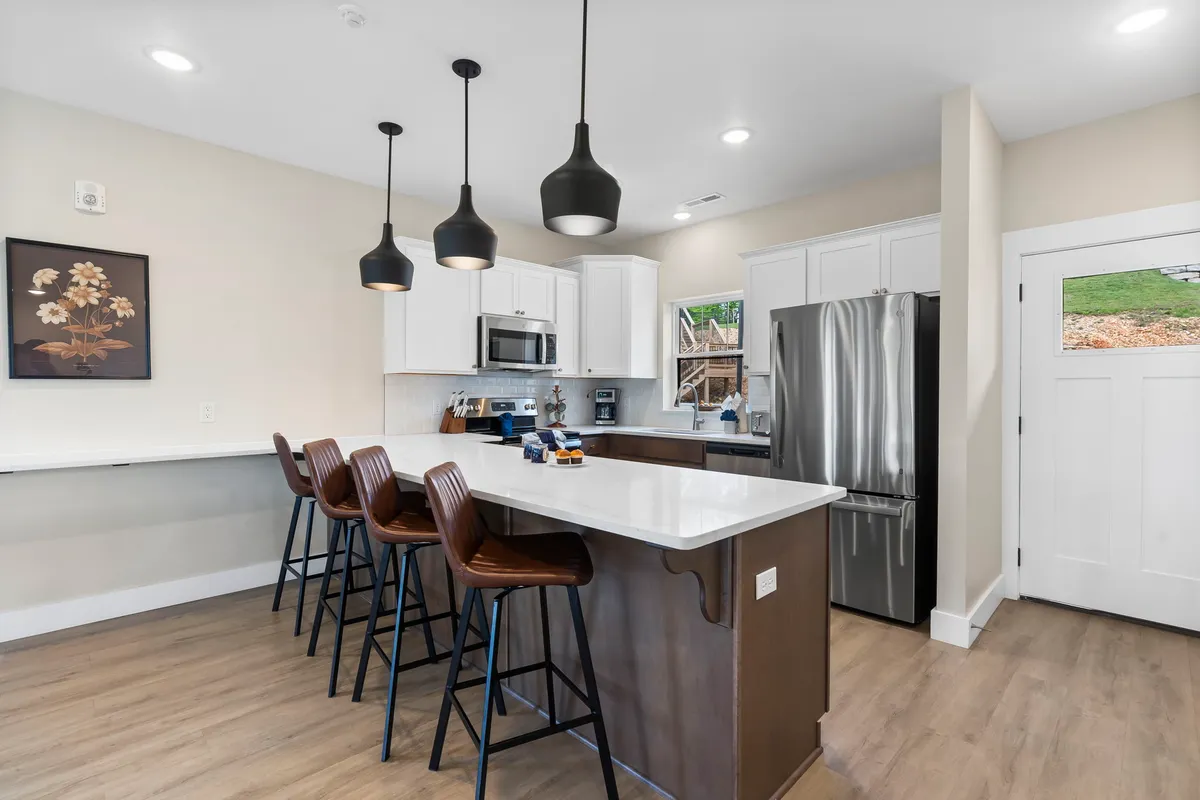 A stylish kitchen featuring a central island with bar stools, pendant lighting, stainless steel appliances, and a window offering a peek outdoors. The countertop displays mugs, hinting at casual coffee moments, while the inviting setup suggests a space for shared cooking and conversation.